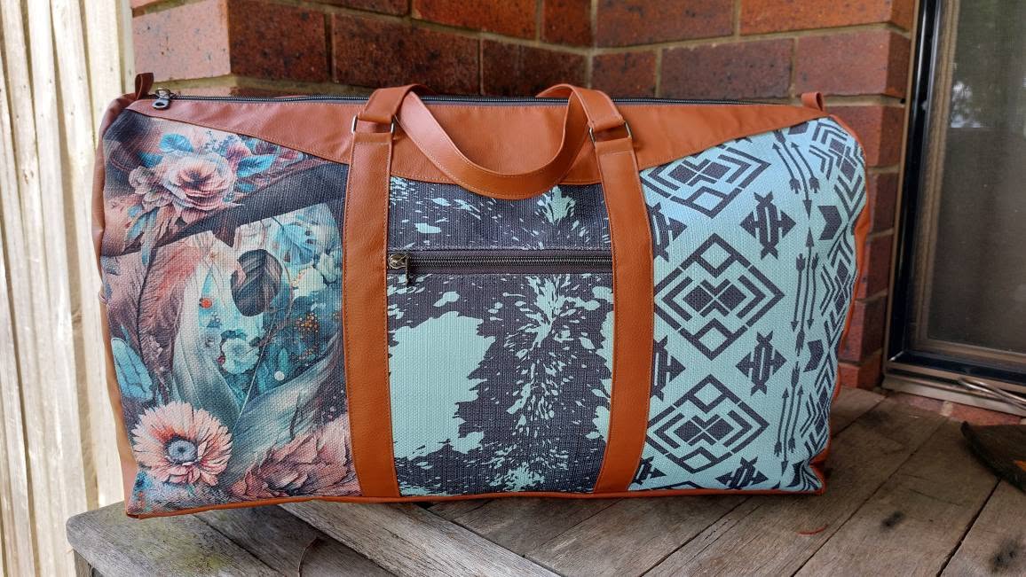Multicolored patterned bag with brown handles on a wooden surface against a brick wall.